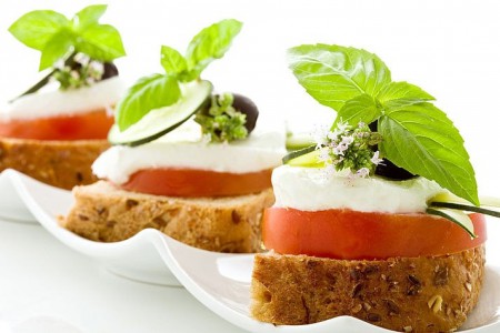 Rodajas de variedad de pan es integrales con montaje de tomate, queso blando, zapallito glaseado y cierre con aceituna y hoja de albahaca.